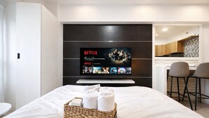 A hotel room with a large flat-screen TV mounted on the wall displaying Netflix content, a neatly made bed with a white comforter, a wicker basket with towels, and a modern kitchen area visible through an open door. - Noirwood Studio | IT Park Pool + Fast WiFi (Cebu City)
