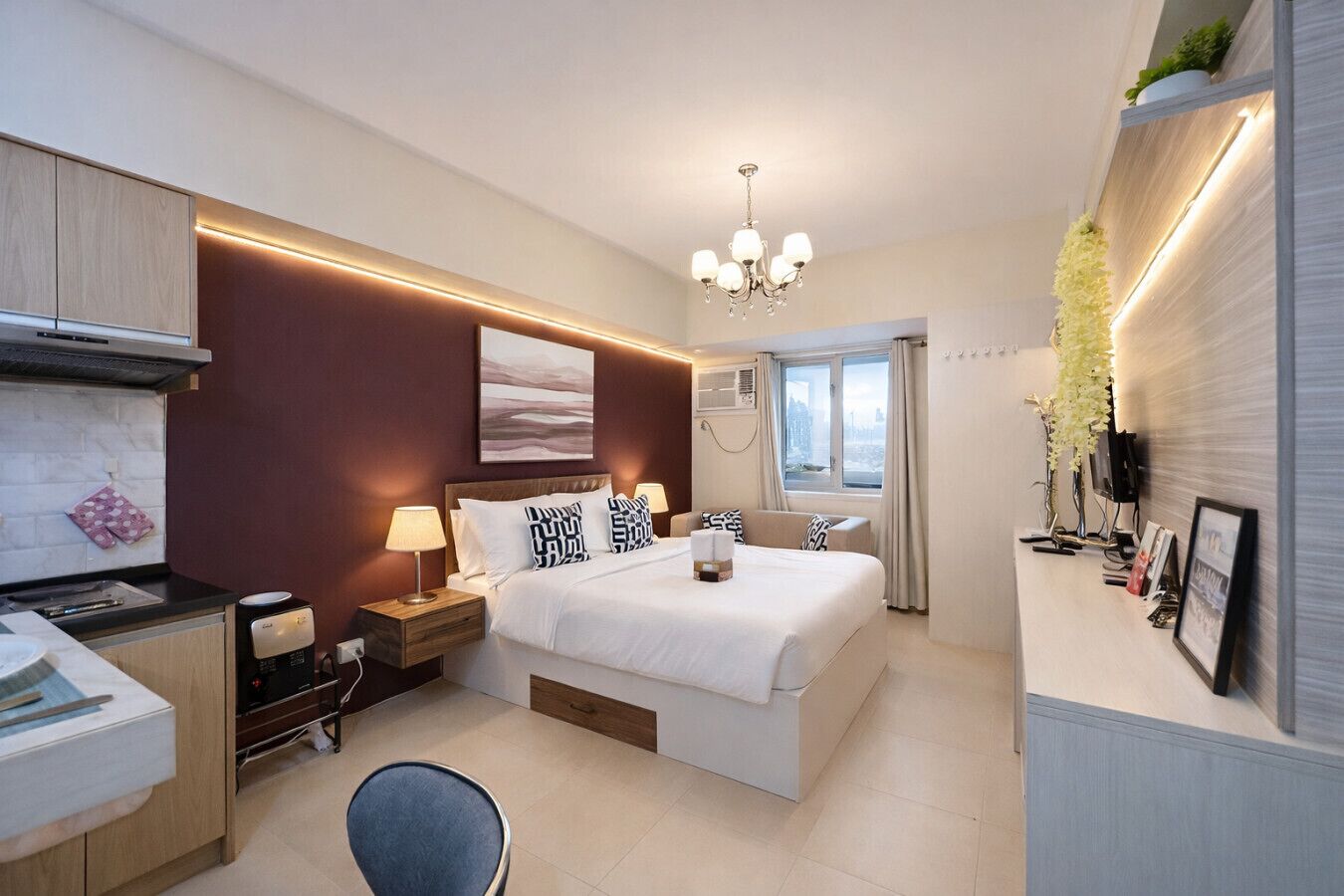 A modern hotel room with a bed, bedside tables, a desk, a chair, a chandelier, and a window with curtains.