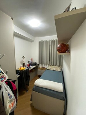 A compact hotel room with a single bed, a desk with a chair, a small shelf with a basketball, and a curtain. - Apartment 68m² | 3 bedrooms (1 suite) | Furnished | 2 parking spaces | Balcony and service area (São Paulo)