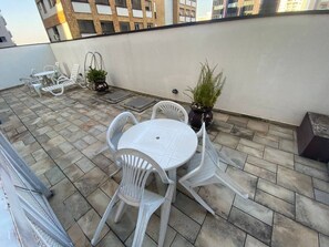 A rooftop terrace with a round table and white plastic chairs, potted plants, and a view of buildings in the background. - Apartment 68m² | 3 bedrooms (1 suite) | Furnished | 2 parking spaces | Balcony and service area (São Paulo)