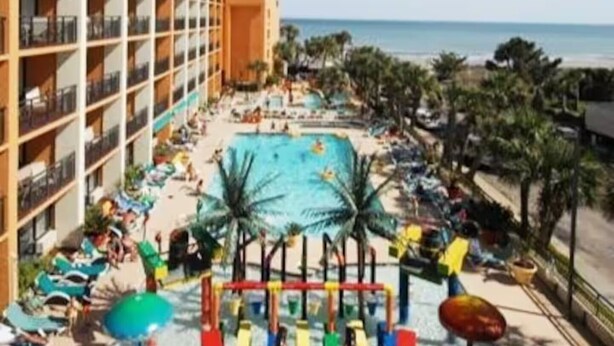 A resort pool area with a view of the ocean, multiple levels of balconies, and palm trees. - Relaxing 1 bedroom oceanfront. (Myrtle Beach)