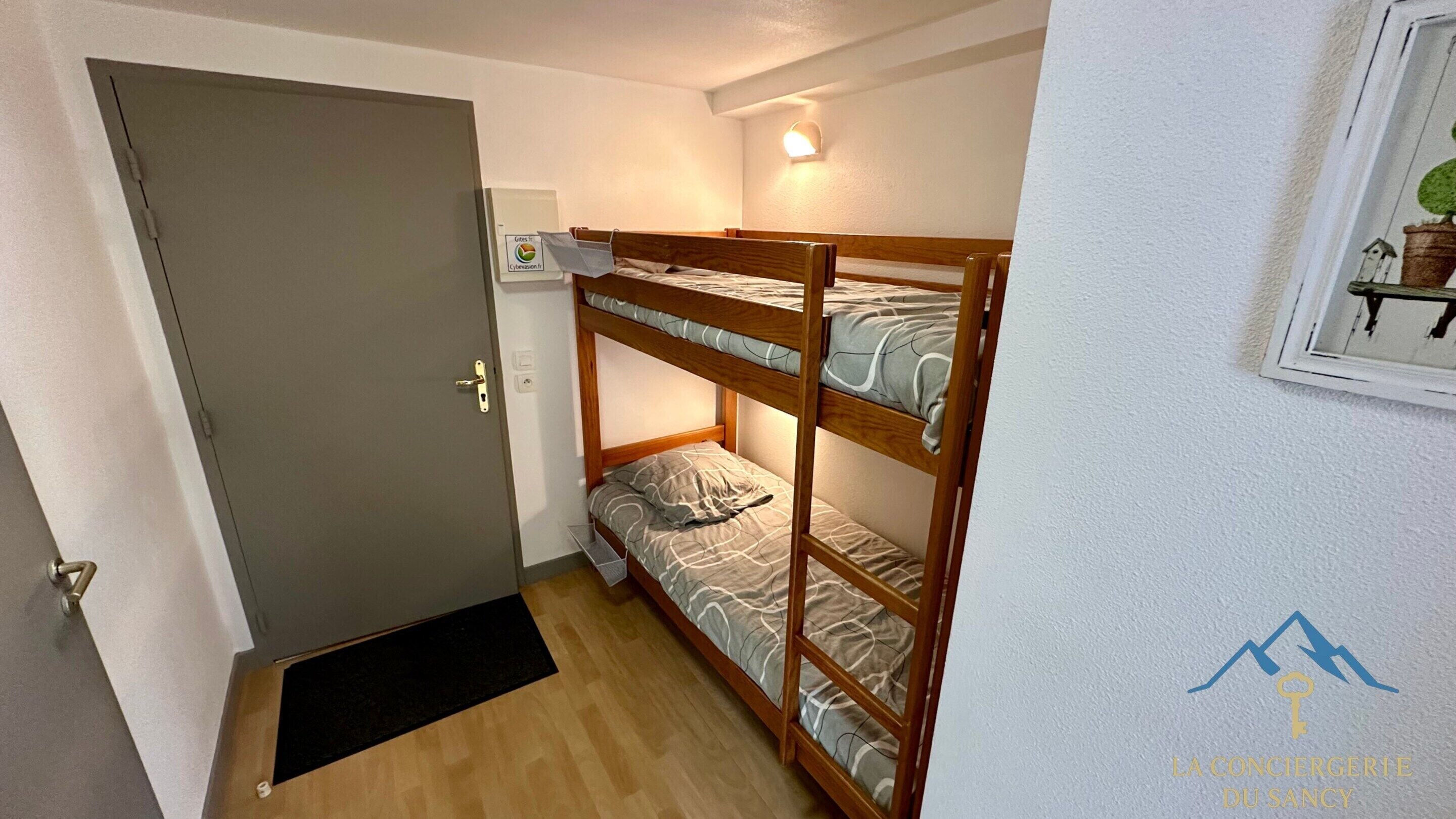 Ein Zimmer mit einem Etagenbett, einer Tür, einer Wandleuchte und einem kleinen Fenster mit einer Pflanze.
