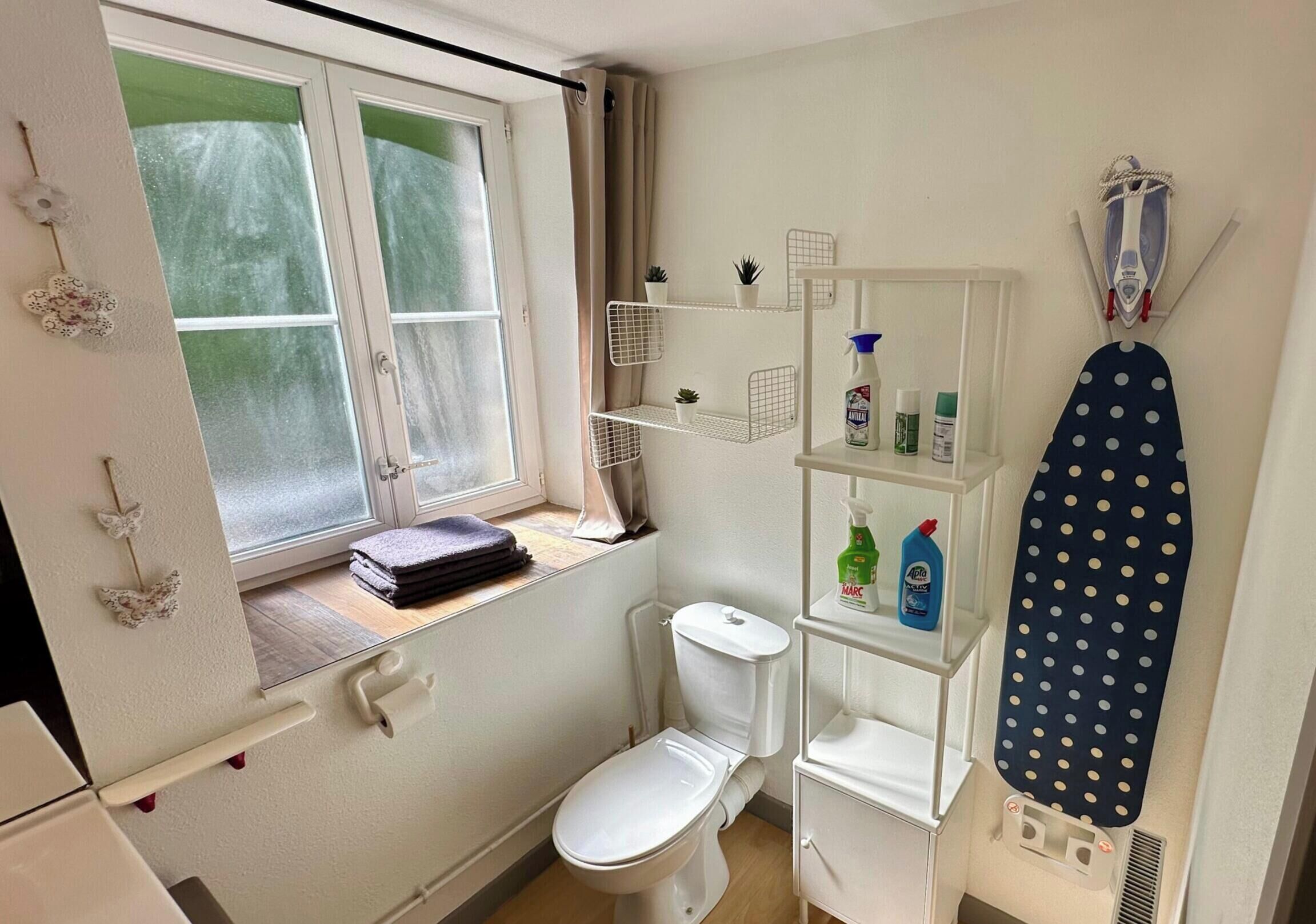 A compact bathroom with a toilet, a window with curtains, a shelf with cleaning supplies, and a polka-dotted ironing board.