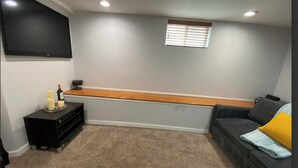 Ein Wohnzimmer mit einem an der Wand montierten Flachbildfernseher, einer Holzbank, einem schwarzen Sofa mit gelbem Kissen und einem kleinen Tisch mit Kerzen und einer Flasche.