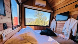 Ein Schlafzimmer einer Holzhütte mit Bett, Kissen, einem Fernseher und einem Fenster mit Blick auf Bäume und Hügel.