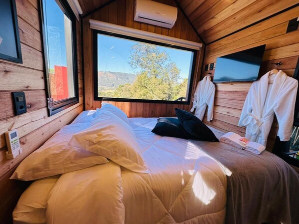 A wooden cabin bedroom with a bed, pillows, a TV, and a window with a view of trees and hills. - Chalé Harmonia da Floresta (Urubici)