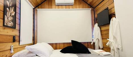 Een slaapkamer met een houten plafond, een bed met wit beddengoed, een zwart kussen en een grijze deken. Aan de muur hangt een whiteboard en erboven een flatscreen-tv.