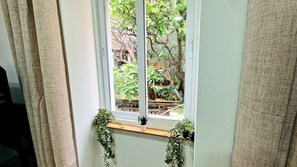 Une fenêtre donnant sur un jardin, avec des rideaux de chaque côté et une petite étagère où sont posées des plantes en pot.