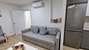 Un salon moderne avec un canapé gris, une petite table sur laquelle se trouve un panier, et un réfrigérateur en acier inoxydable.