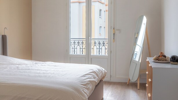Un dormitorio con cama, cómoda, espejo y ventana con vista a edificios.