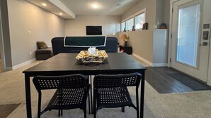 A modern living room with a dark wooden table, two black chairs, a sofa with a green throw, and a TV mounted on the wall. - Family friendly, super clean & Cozy Walkout basement suite with all amentities (Calgary)