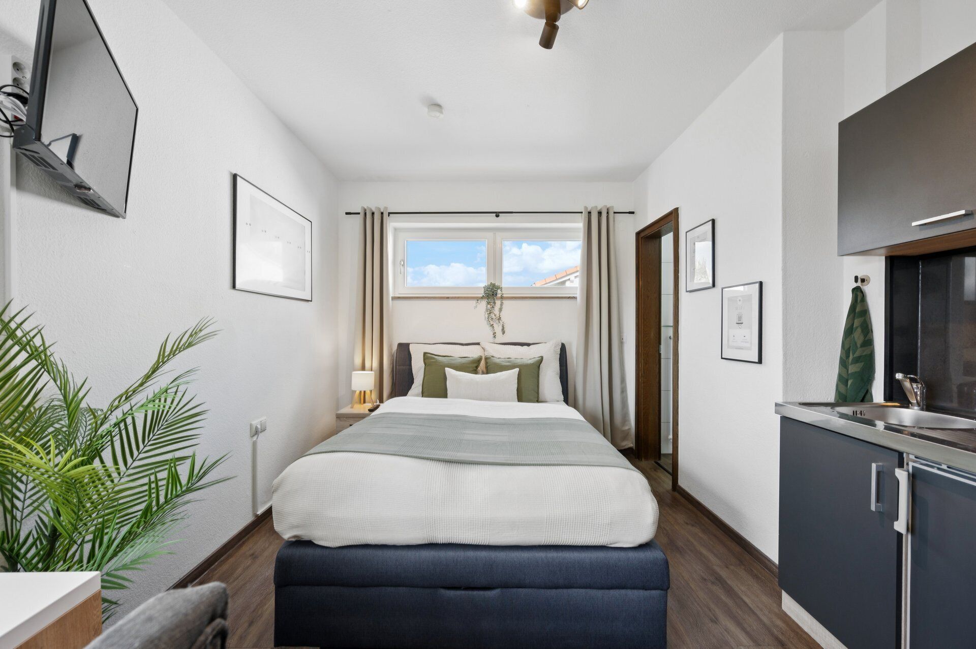 A modern hotel room with a bed, bedside table, lamp, and a view of the sky through a window.