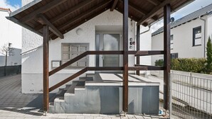 Un bâtiment à l’extérieur blanc, avec un escalier en bois menant à une porte, et une clôture en métal au premier plan.