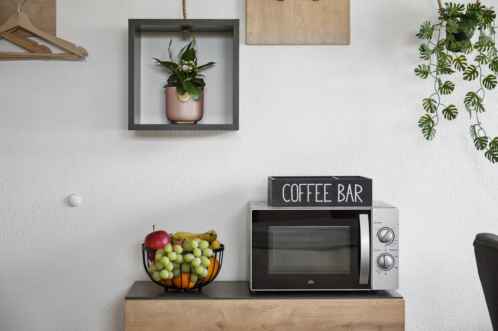 Eine Mikrowelle mit einem 'Coffee Bar' Schild, eine Obstschale, eine hängende Pflanze und ein Holzaufhänger an der Wand.