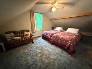 Un dormitorio con techo inclinado, ventilador de techo, ventana con cortinas, cama con cubrecama estampado y sofá con cojines decorativos.