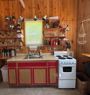 A compact kitchen with a stove, sink, and shelves filled with kitchenware. - Loon-R Landing on paddle only Daniels Pond in Glover, Vermont (Glover)