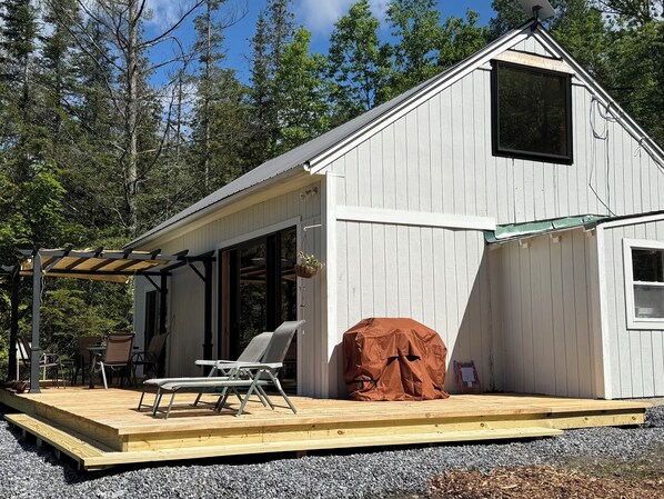 Eine kleine Holzhütte mit einer Terrasse, einem überdachten Grill und einer Veranda mit Stühlen.