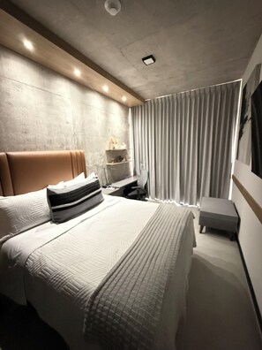 Une chambre d’hôtel moderne, dotée d’un grand lit, d’une table de chevet, d’une lampe et offrant une vue sur l’extérieur à travers des rideaux.