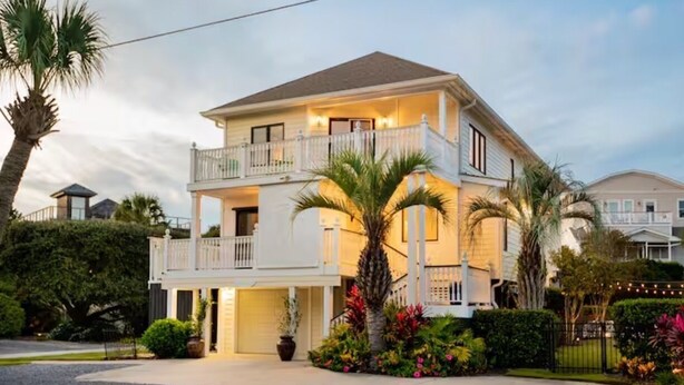 Une maison à deux étages avec un balcon au deuxième étage, entourée de palmiers et d’un jardin bien entretenu.