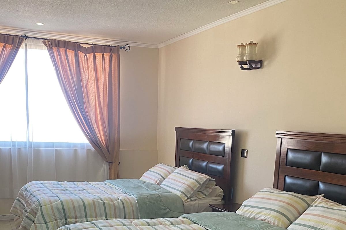 Ein Hotelzimmer mit drei Betten, einem großen Fenster mit Vorhängen, einer Wandleuchte und einem braunen Teppich.
