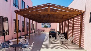 A covered outdoor seating area with tables and chairs, a pergola, and a view of a pink building.