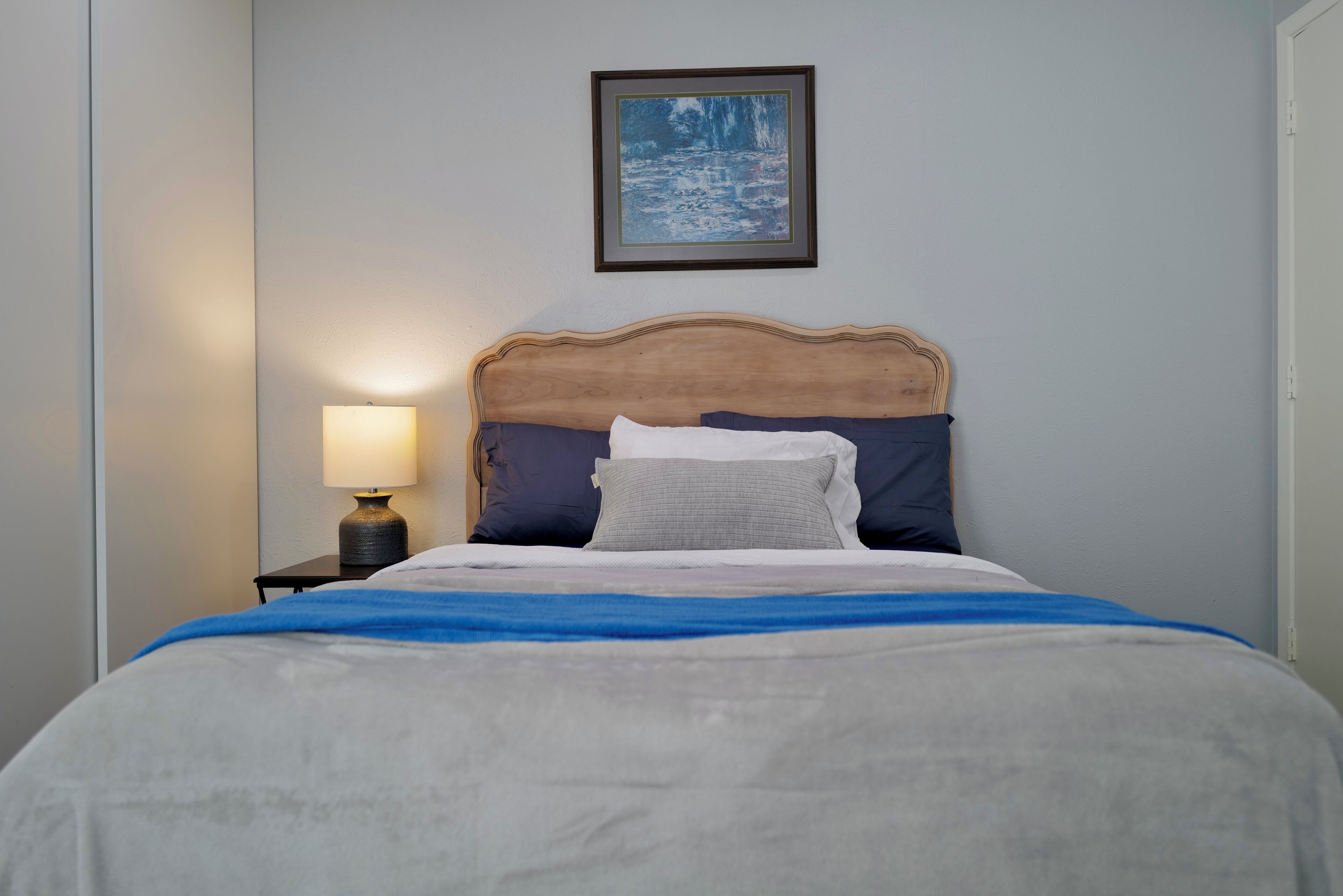 A neatly made bed with a blue blanket, a wooden headboard, and a framed picture on the wall.