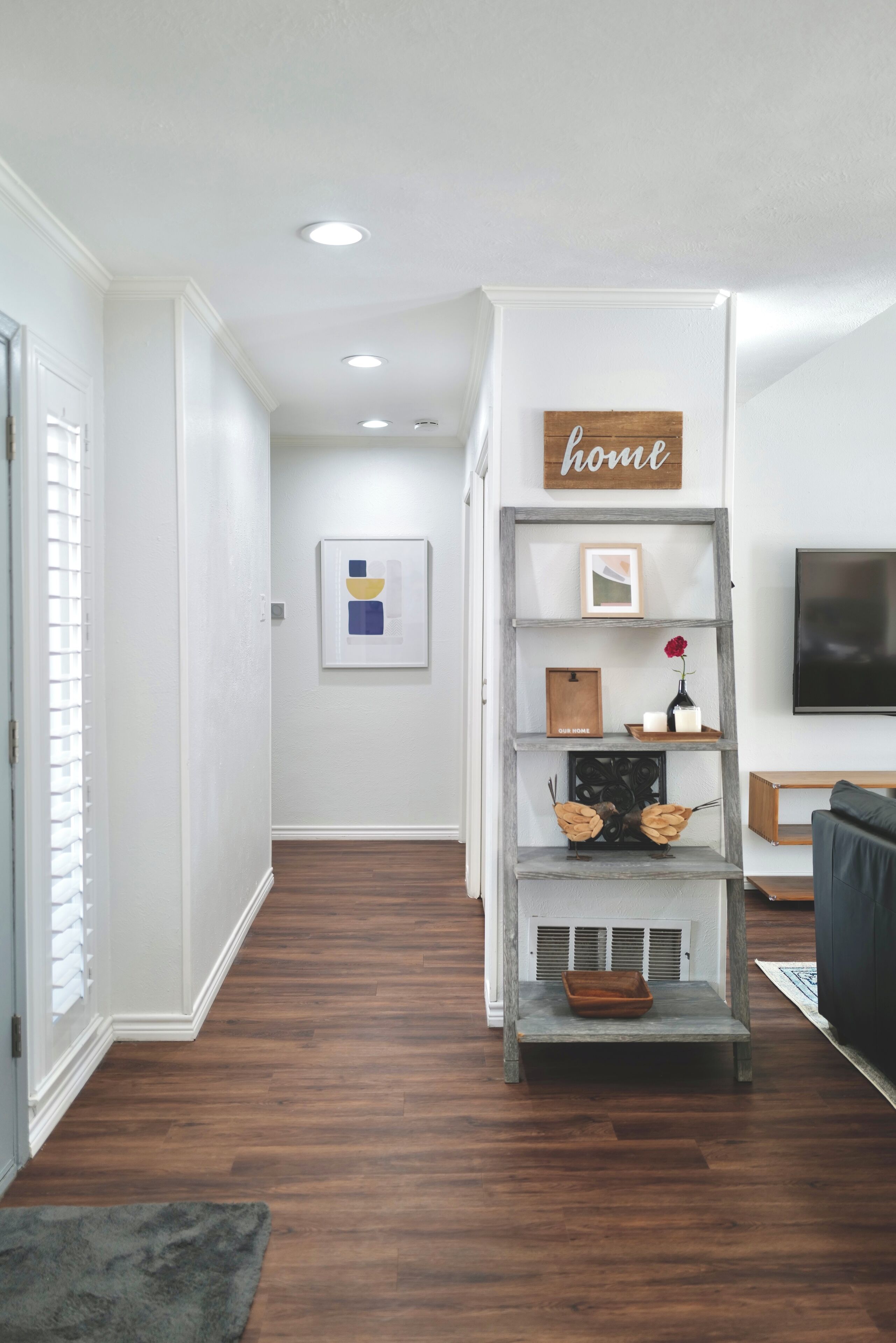 A hallway with a wooden floor, a gray bench, a shelf with decorative items, and a 'home' sign on the wall.