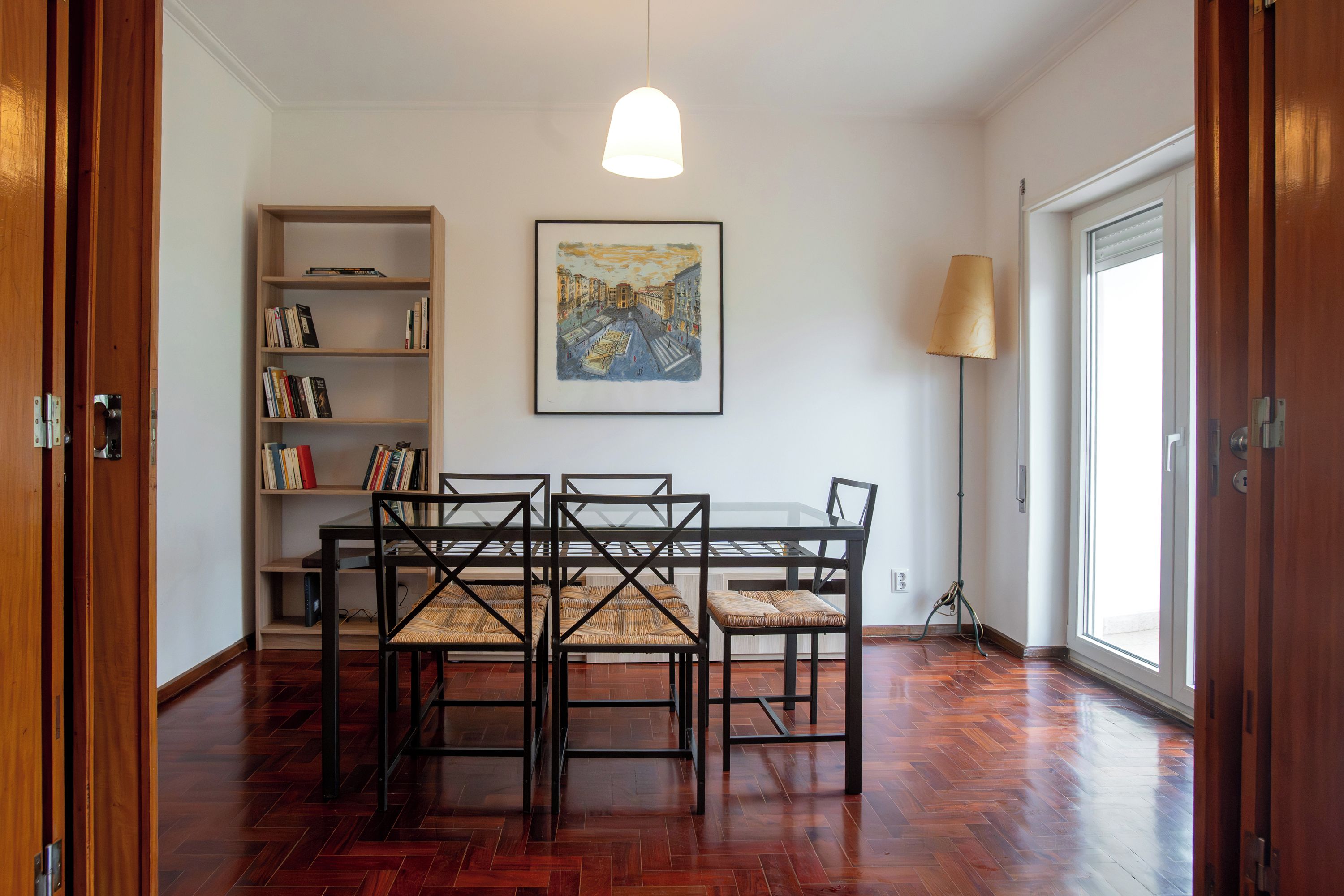A dining area with a table and chairs, a bookshelf, a painting, and a floor lamp.