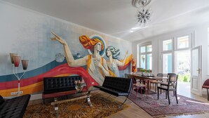 Ein modernes Wohnzimmer mit einer großen abstrakten Wandmalerei, einem schwarzen Sofa, einem Essbereich mit Tisch und Stühlen und einem großen Fenster mit Blick.
