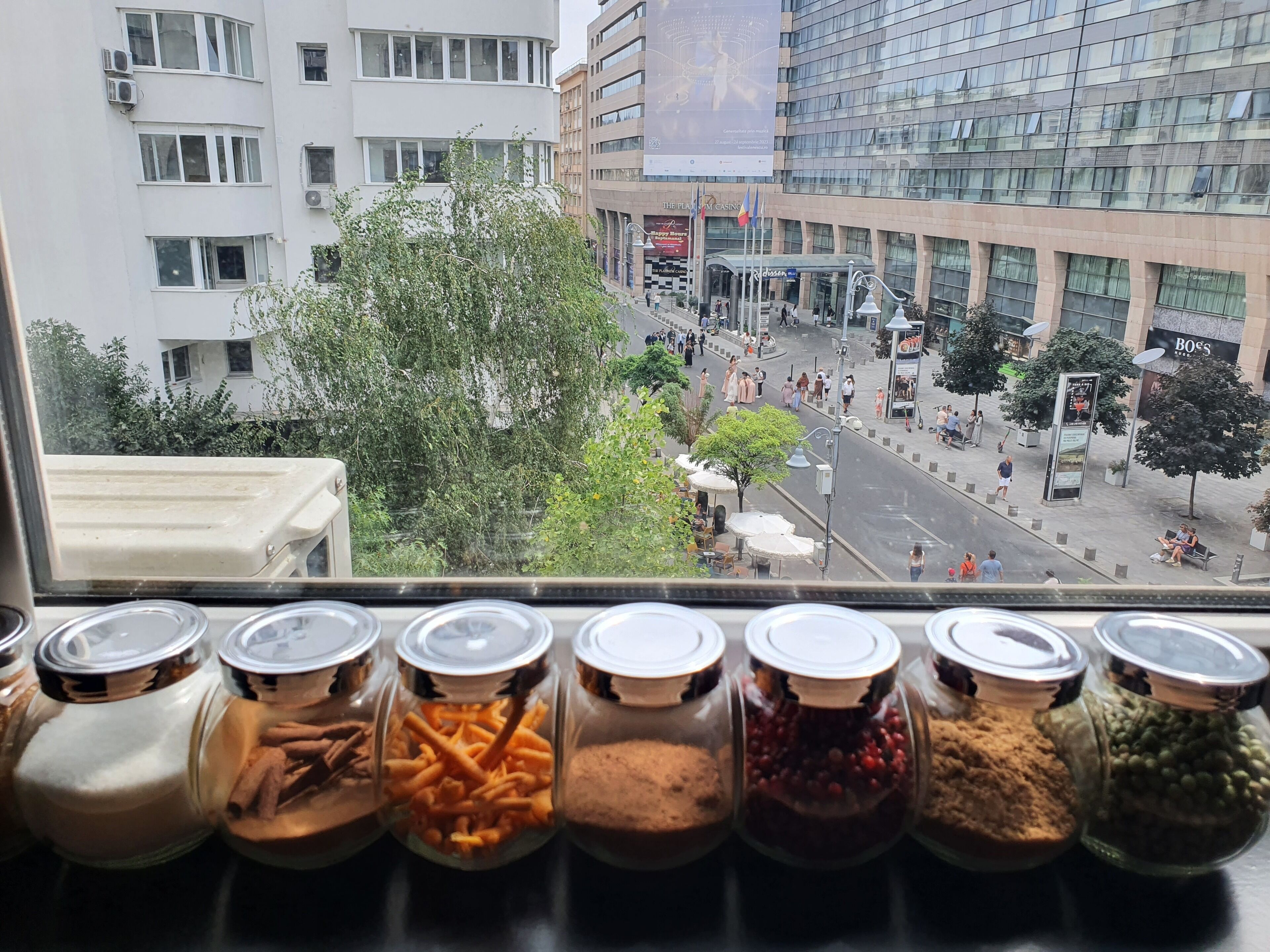 Blick aus einem Fenster auf eine Straße mit Gebäuden, Bäumen und einem Fußgängerbereich.