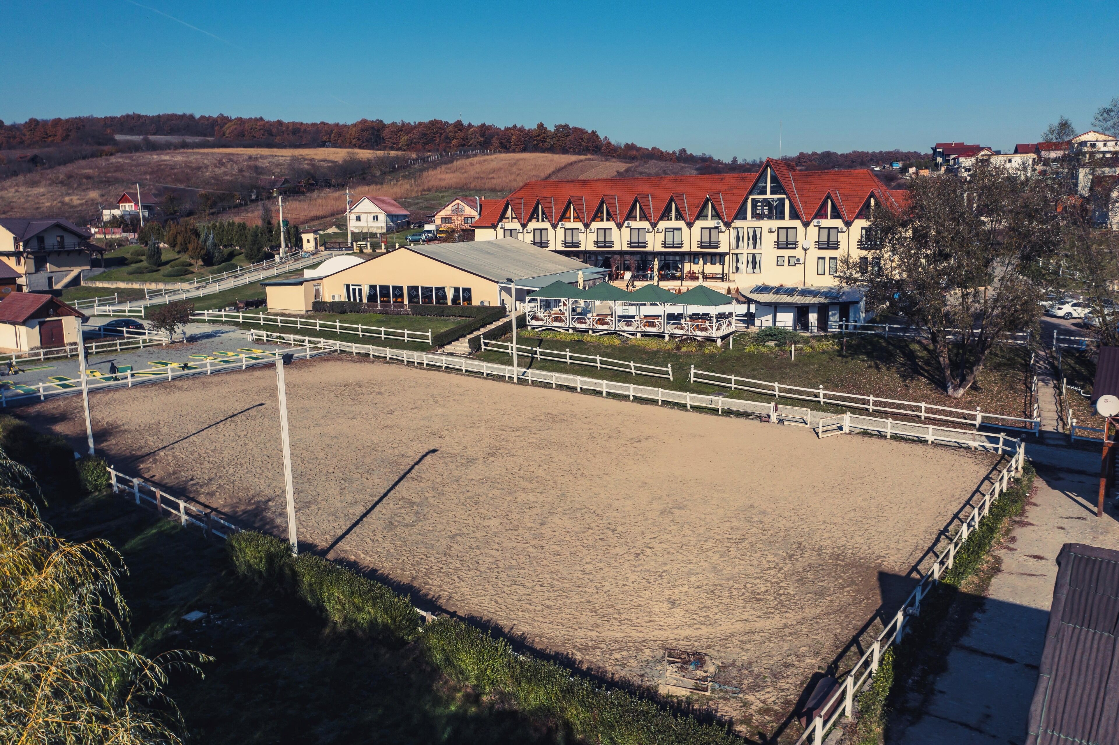 A large equestrian arena with a white fence, surrounded by buildings with red roofs and a hill in the background.