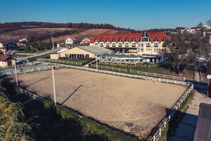 A large equestrian arena with a white fence, surrounded by buildings with red roofs and a hill in the background.