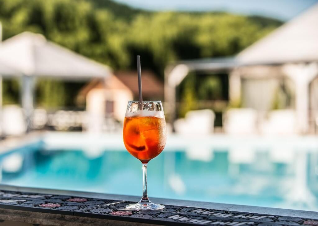 Ein Glas bernsteinfarbenes Getränk auf einem Tisch am Pool, im Hintergrund Pool und Sonnenschirme.