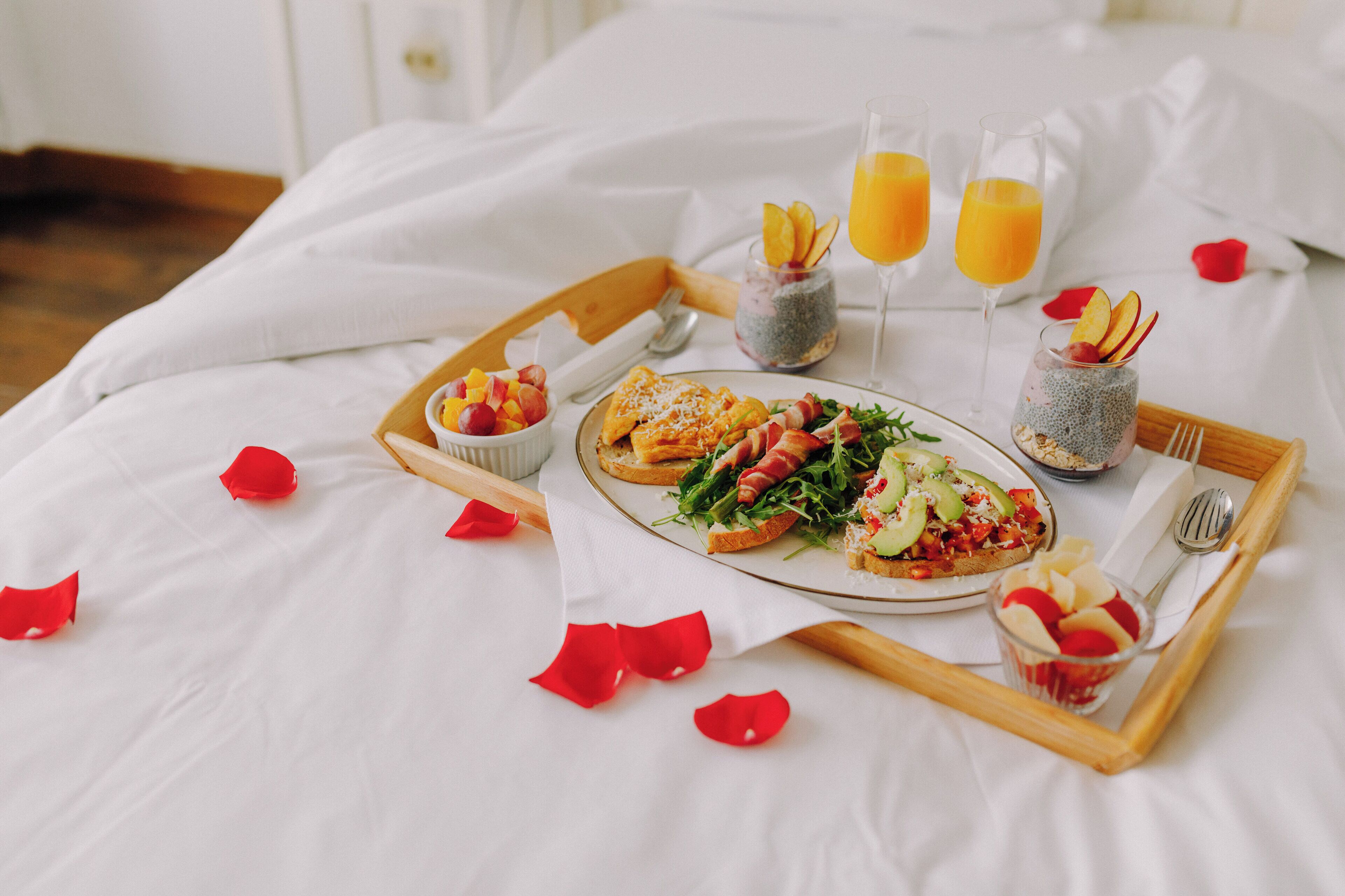 A breakfast tray with toast, fruit, and juice on a bed with white linens and rose petals.