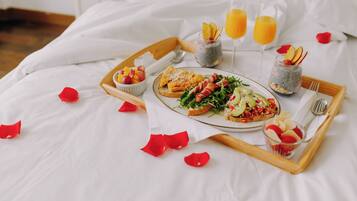 A breakfast tray with toast, fruit, and juice on a bed with white linens and rose petals.