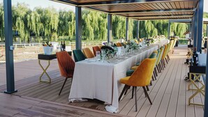 A covered outdoor dining area with a long table set for a meal, surrounded by colorful chairs, and a view of a natural landscape.