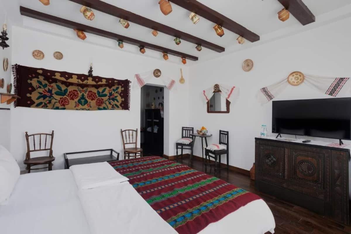 A room with a bed, two chairs, a small table, a television, and a decorative wall hanging.