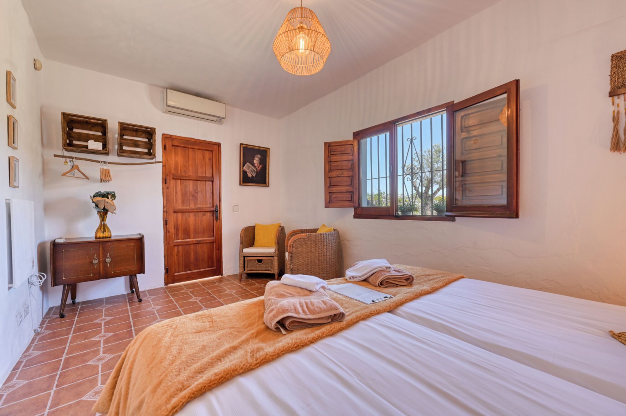 A bedroom with a bed, a wooden wardrobe, a wicker chair, a hanging light fixture, and a window with shutters.