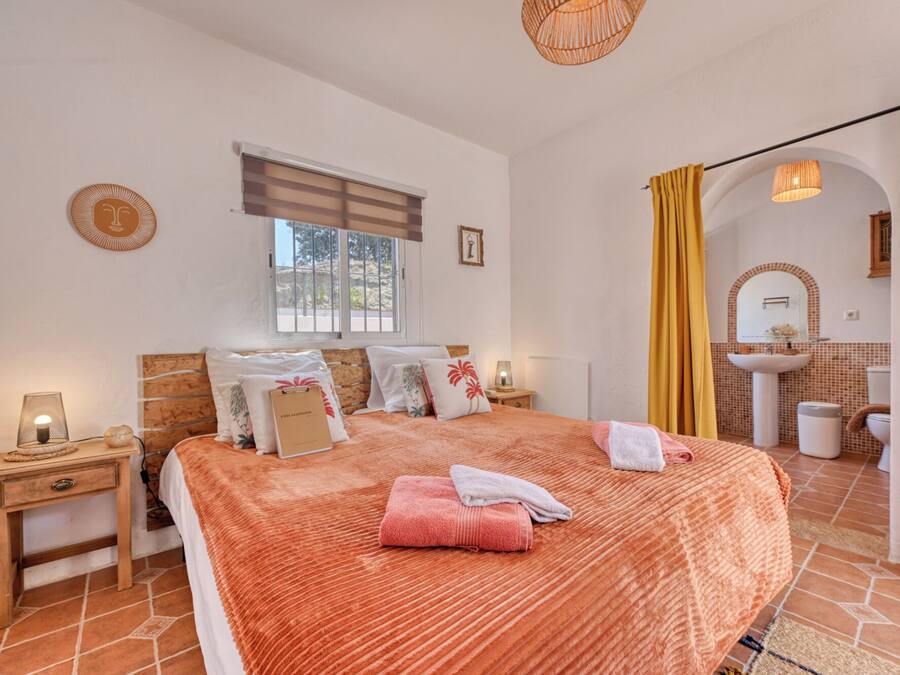 Un dormitorio con cama, mesitas de noche, una ventana, un baño con lavabo y espejo, y una cortina amarilla.
