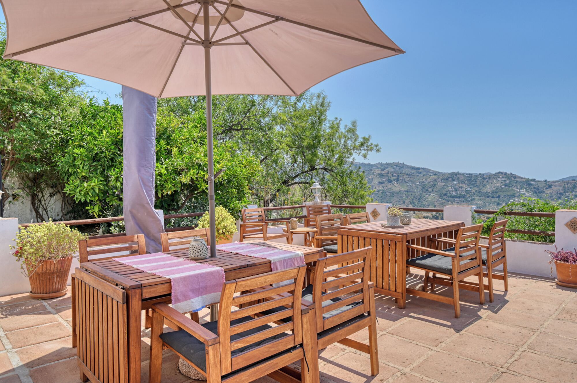Un patio al aire libre con mobiliario de madera, un paraguas grande y vista a una ladera.