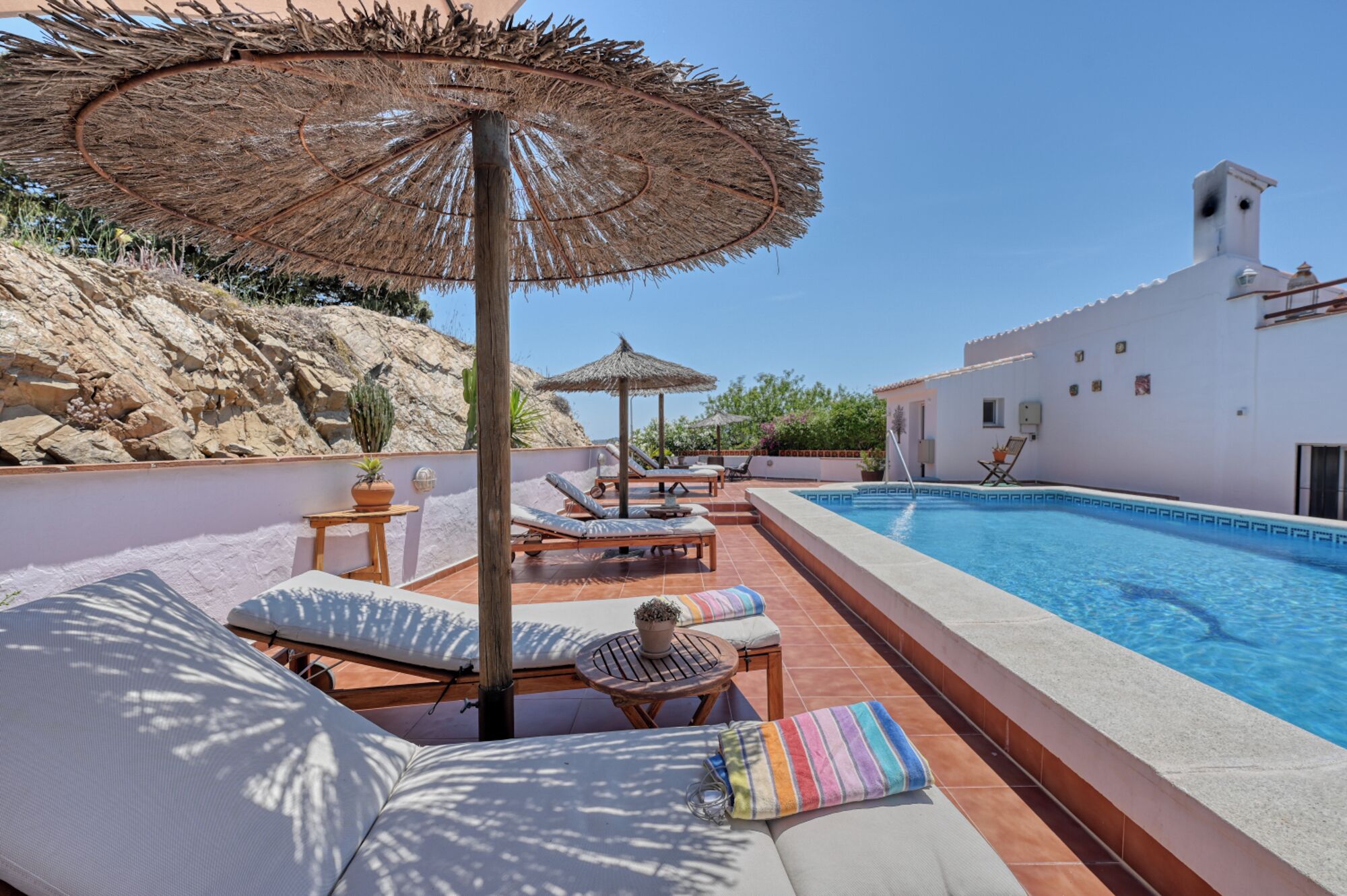 A poolside area with lounge chairs, a thatched umbrella, and a view of a white building with a tiled roof.