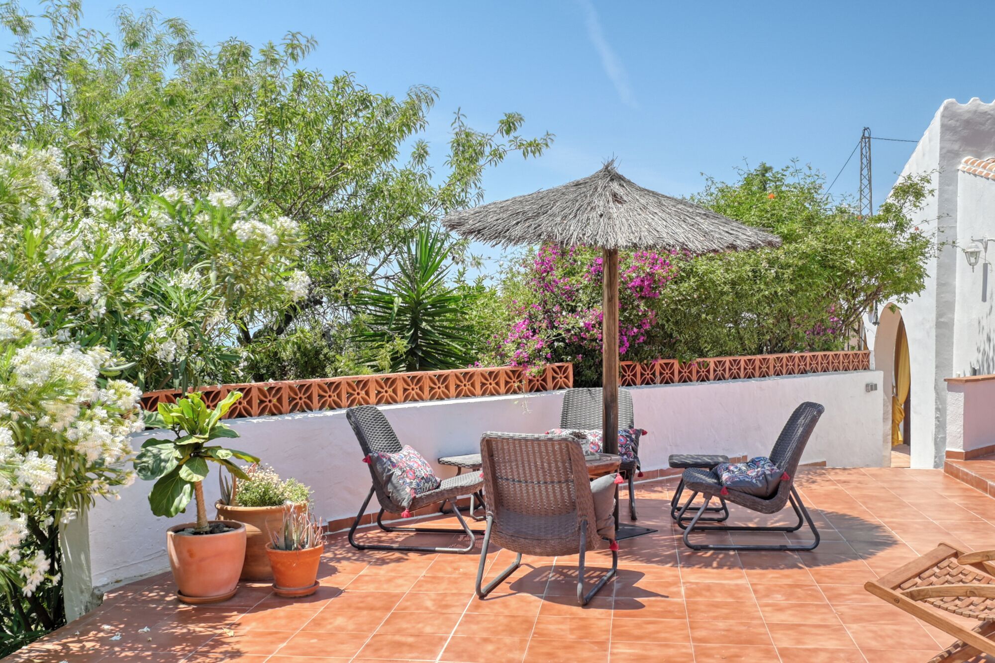 Un patio con dos sillas de mimbre, una mesita y un paraguas de paja. Hay plantas en macetas y una cerca decorativa.