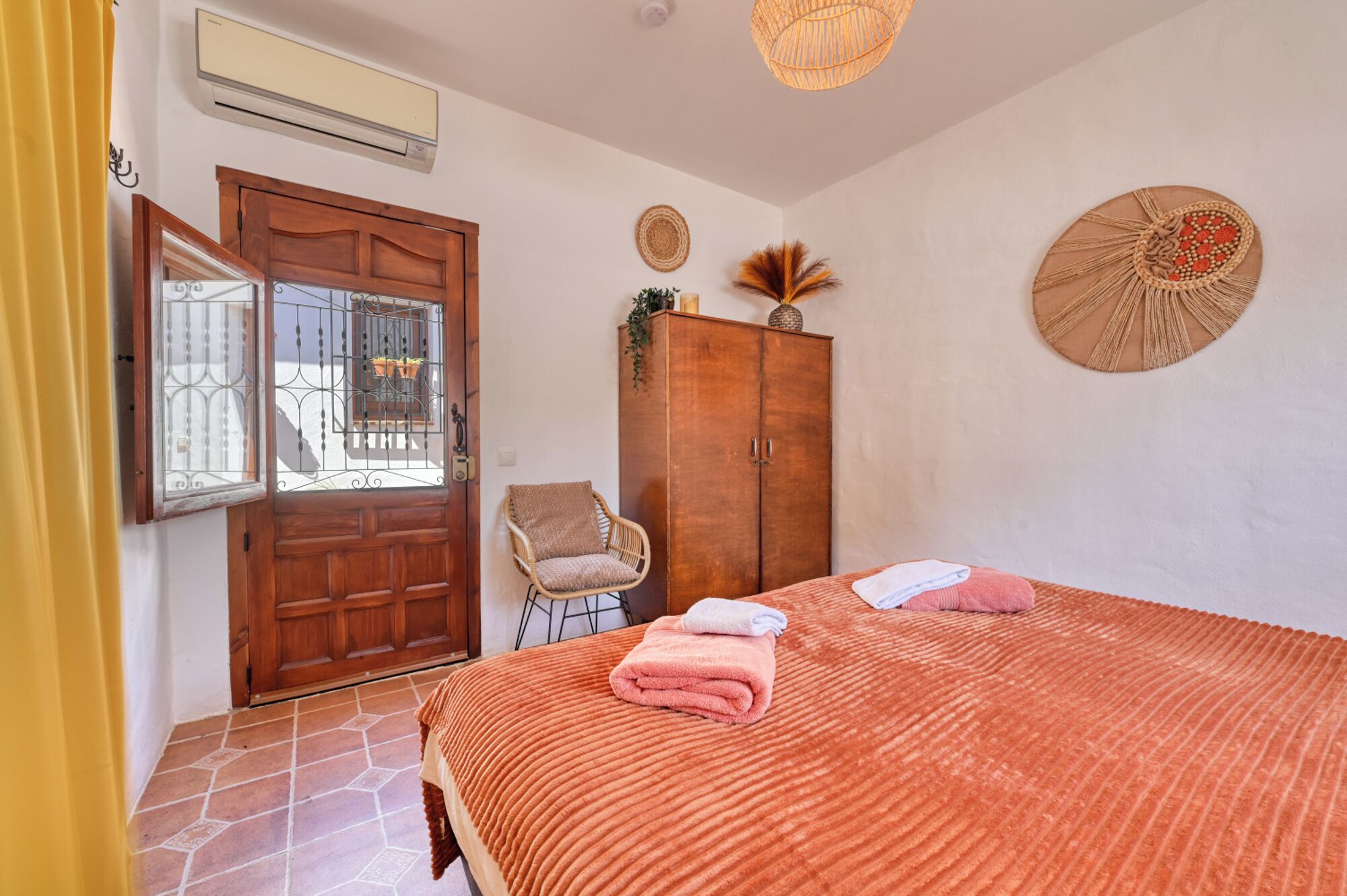 A bedroom with a bed, a wooden wardrobe, a wicker chair, a wall-mounted macrame decoration, and a window with a metal grill.