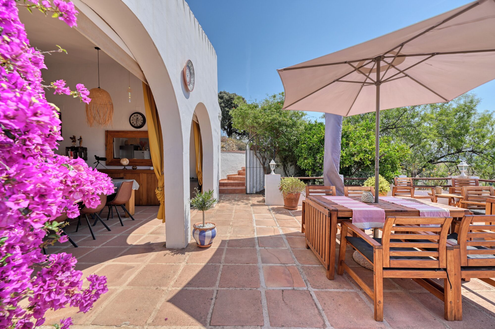 An outdoor patio with wooden tables and chairs, a large umbrella, and a view of the surrounding greenery.
