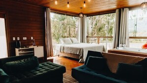 Una habitación acogedora con cama, dos sofás, una cafetera y vistas a árboles a través de amplios ventanales.