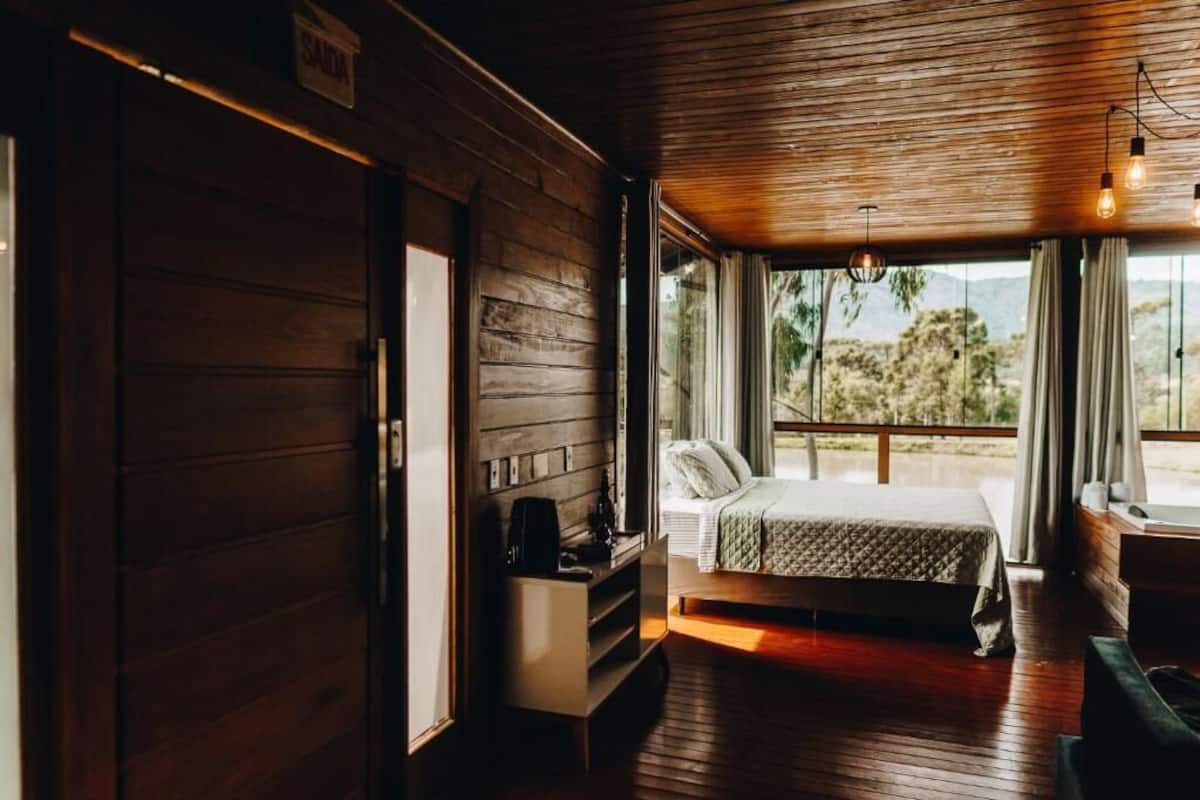 A wooden room with a bed, a small table, and a window with a view of trees.