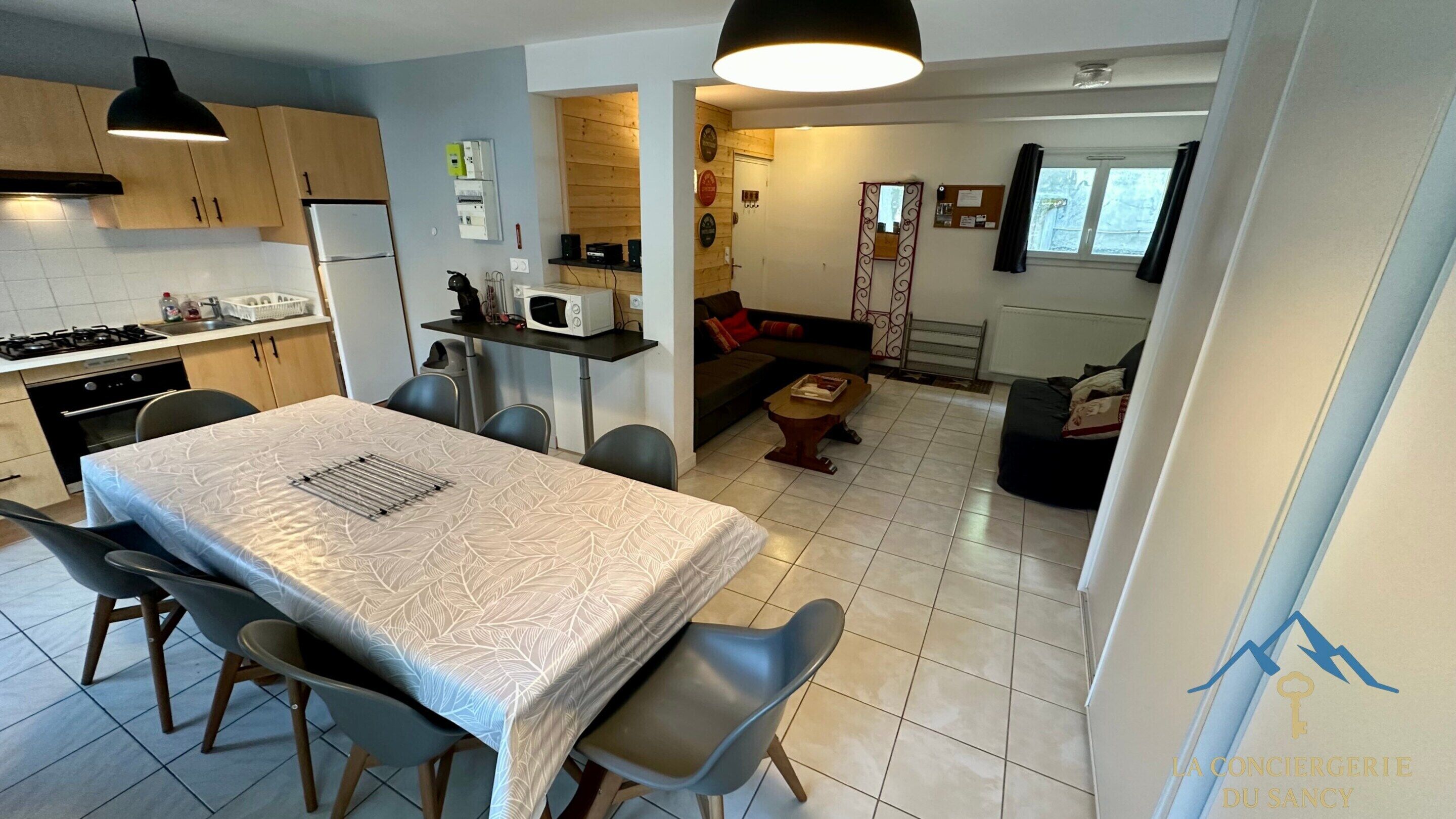 A modern kitchen with a dining area, a living room with a sofa and a coffee table, and a view of the outside through a window.