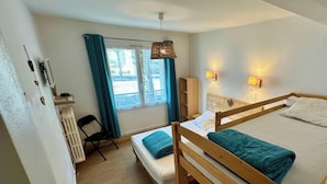 Un dormitorio con dos camas, una ventana con cortinas, un radiador, una silla y una estantería con una lámpara.