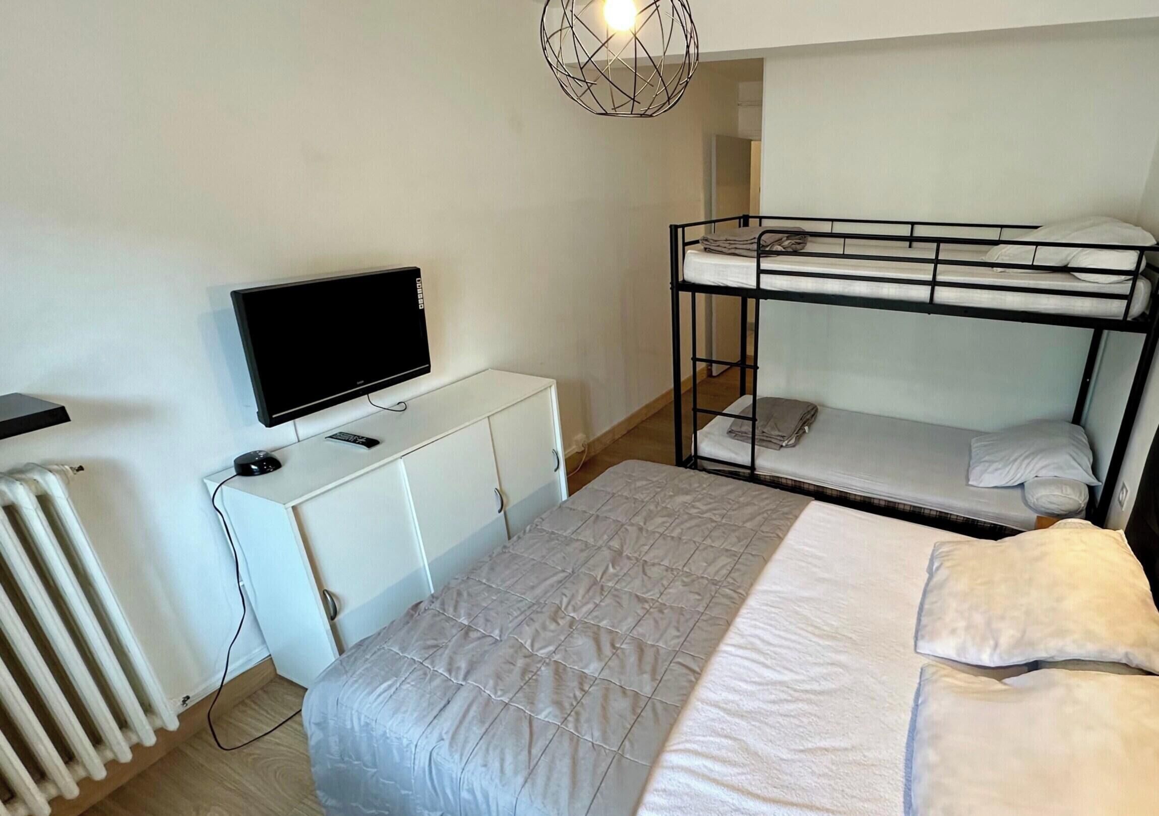 Une chambre avec un lit superposé, une télévision posée sur un meuble blanc, un radiateur et un luminaire suspendu.