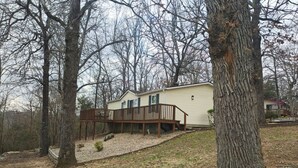 A single-story mobile home with a wooden deck and a small porch, surrounded by trees and a grassy area. - Norfork Lakeview Oasis House #12 at Teal Point Resort (Mountain Home)
